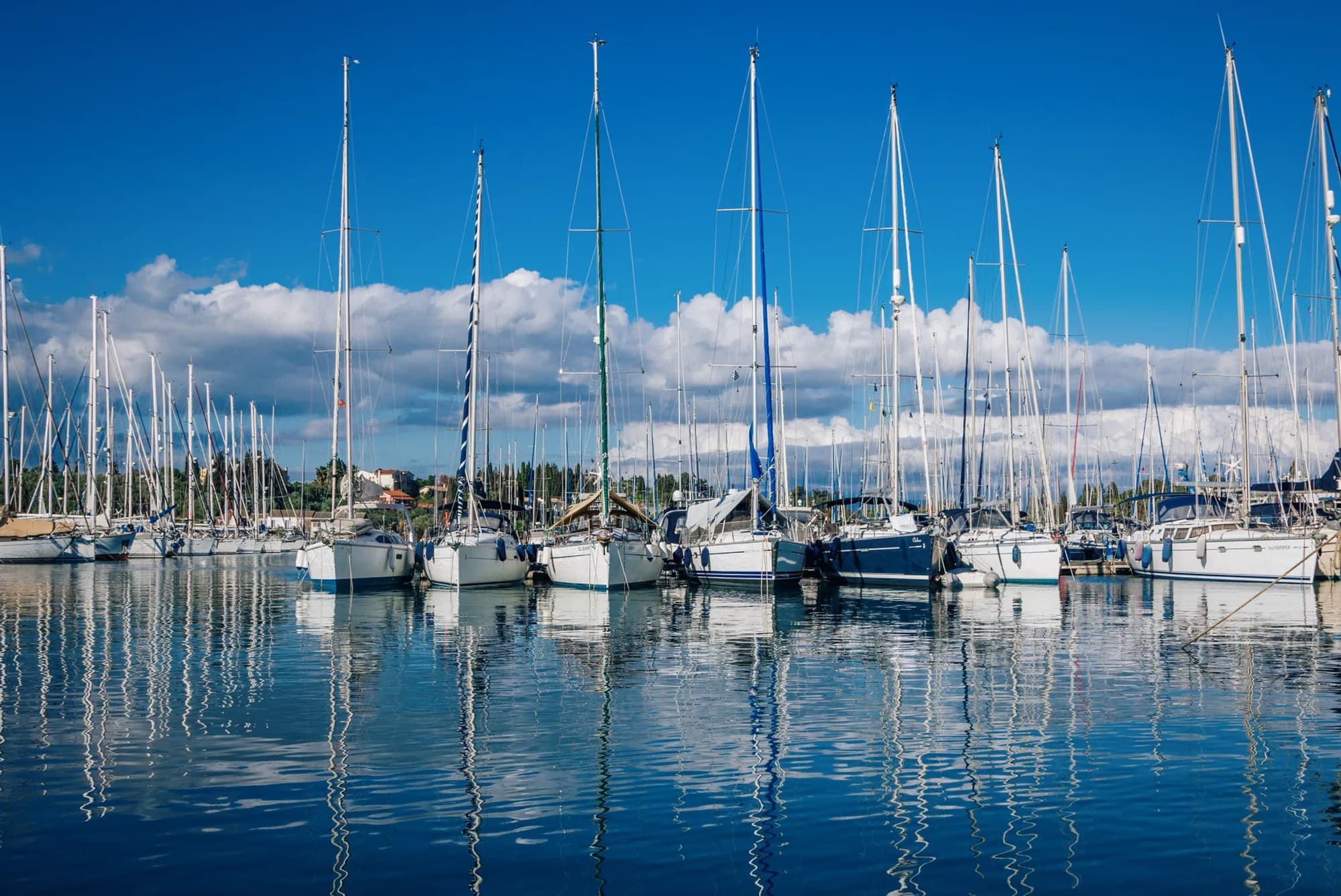 Seilbåter fortøyd på rekke i en marina, med master som speiler seg i det rolige vannet.